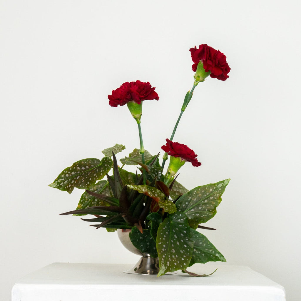 Small floral arrangement with red flowers and green leaves on a white pedestal against a white background