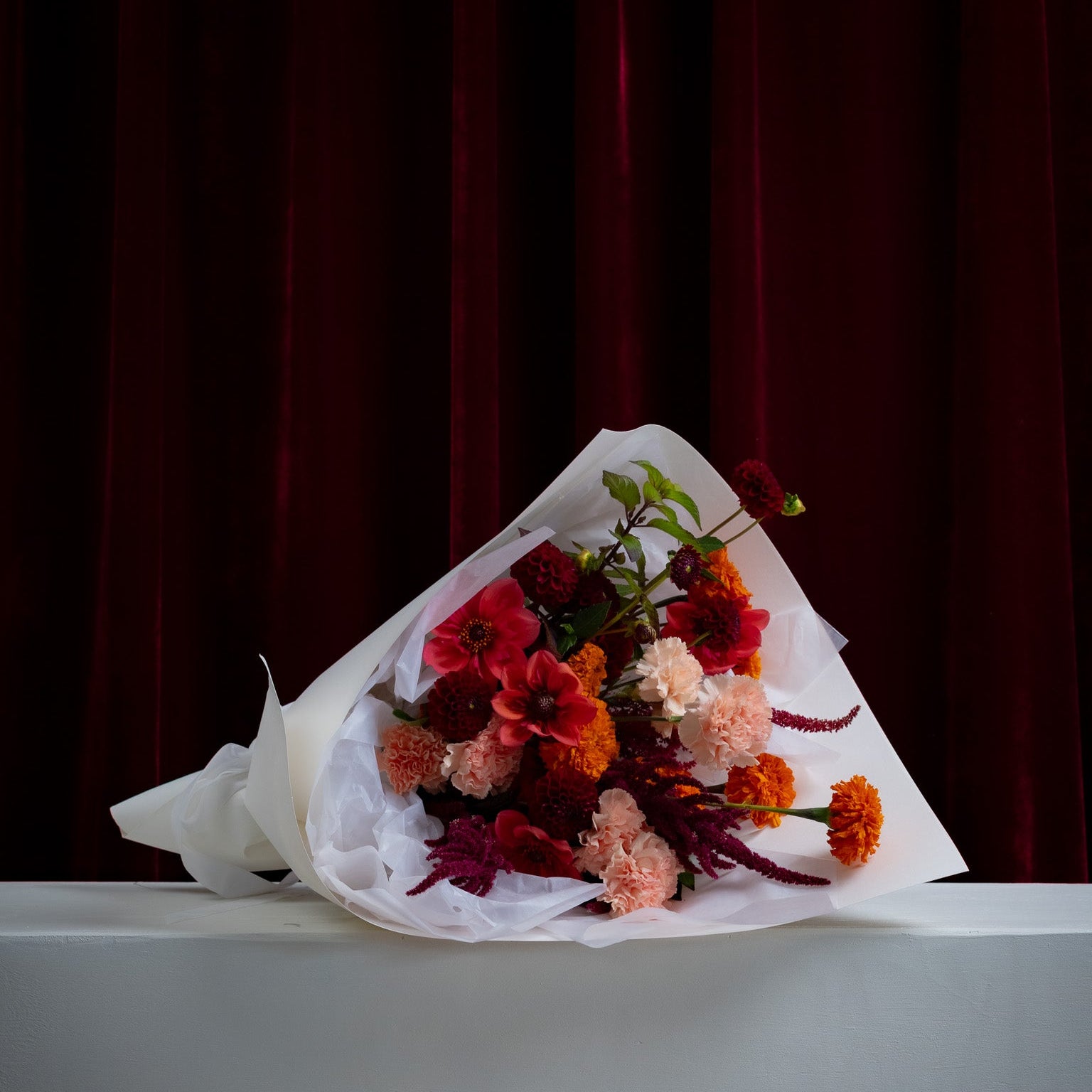 Bouquet of flowers wrapped in white paper on a gray surface with a dark red curtain background
